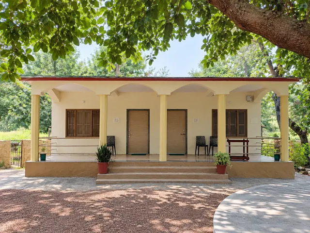 Single-storey cream bungalow with a pillared veranda, steps and potted plants under shady trees