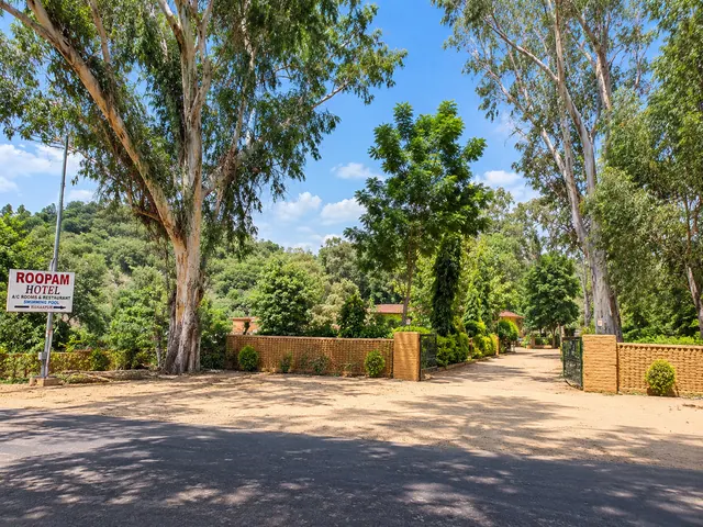 Roadside entrance to Roopam Resort with a signboard, brick boundary wall and gate surrounded by tall trees