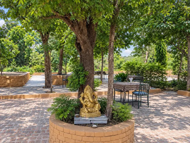 Golden Ganesha statue in a round brick planter beneath a tree, with outdoor tables on the paved garden behind
