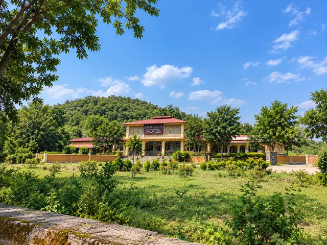 Roopam Resort building seen across a green meadow with forested hills and a blue sky behind