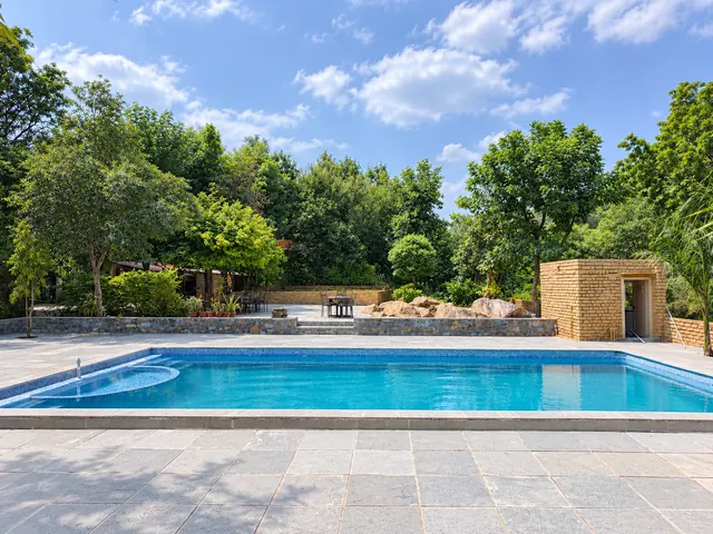 Rectangular swimming pool with a stone sundeck, natural boulders and tall trees against a blue sky at Roopam Resort