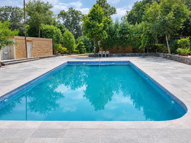 Clear blue swimming pool reflecting the surrounding trees, with a wide stone deck and a small brick building