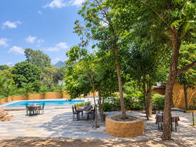 Outdoor tables and chairs beside the swimming pool in the shade of tall trees