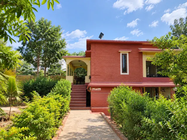 Red-painted two-storey guesthouse with an arched porch and steps, surrounded by shrubs beside a garden path